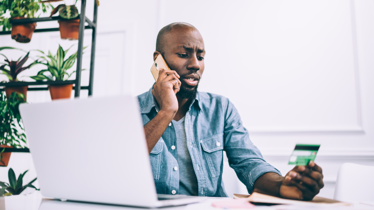 an African American guy in a casual outfit talks on a phone and reads a credit card number while sitting at a cafe table and working