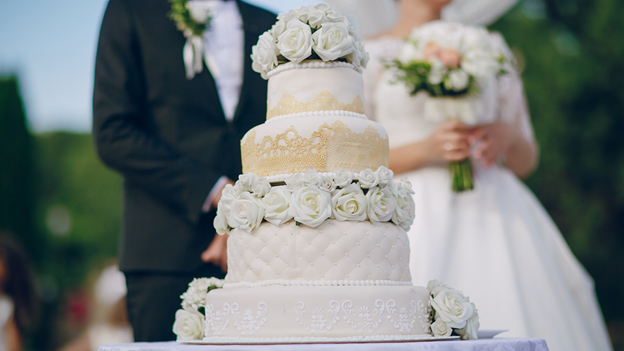 a bride and groom stands behind a four-layer wedding cake decorated with flowers