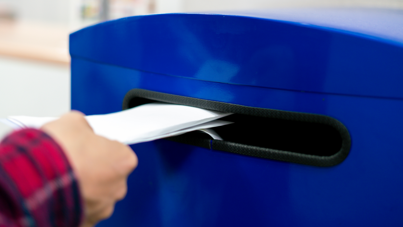 a close-up of a hand dropping a letter and documents in the blue postbox at the post office