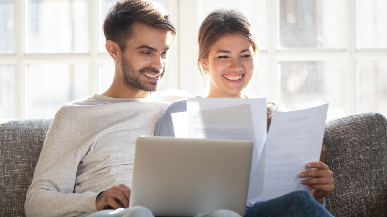 a couple sits on couch and looks at paperwork and a laptop computer screen