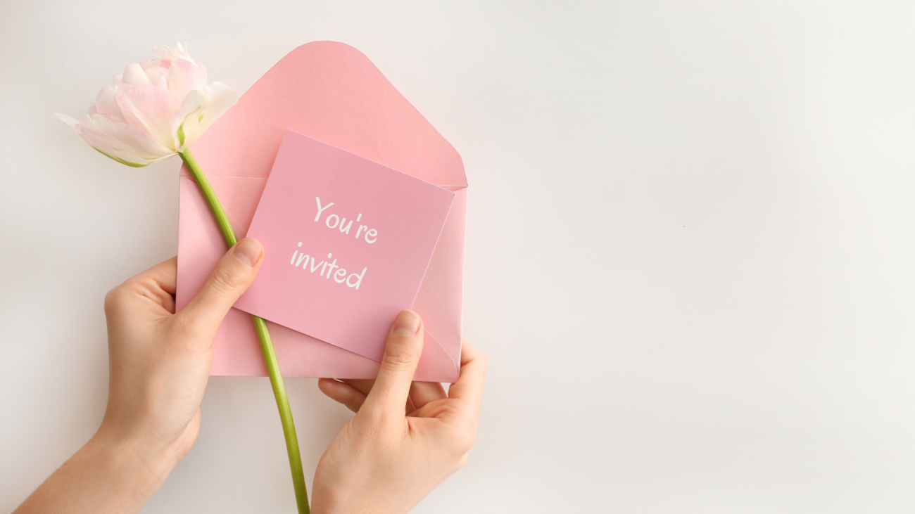 a close-up of a woman’s hands holding a white flower, a pink envelope, and a pink card with the words, “you’re invited” written on the front