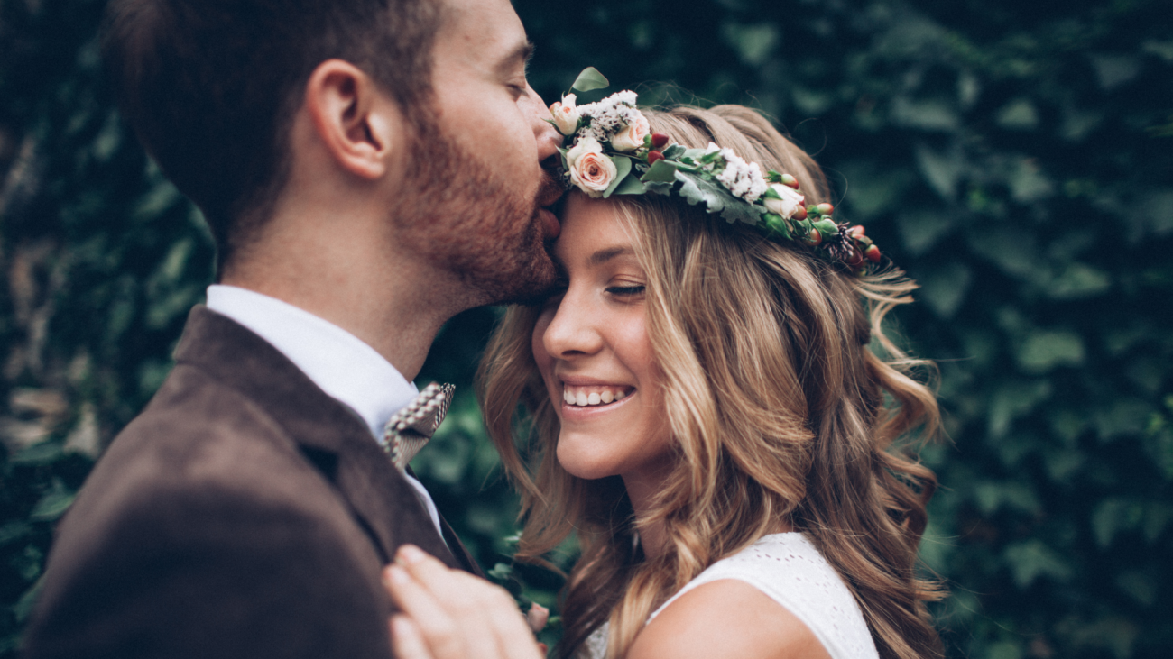 a groom kissing his bride, who is wearing a flower crown and has her eyes closed, on her forehead on their wedding day