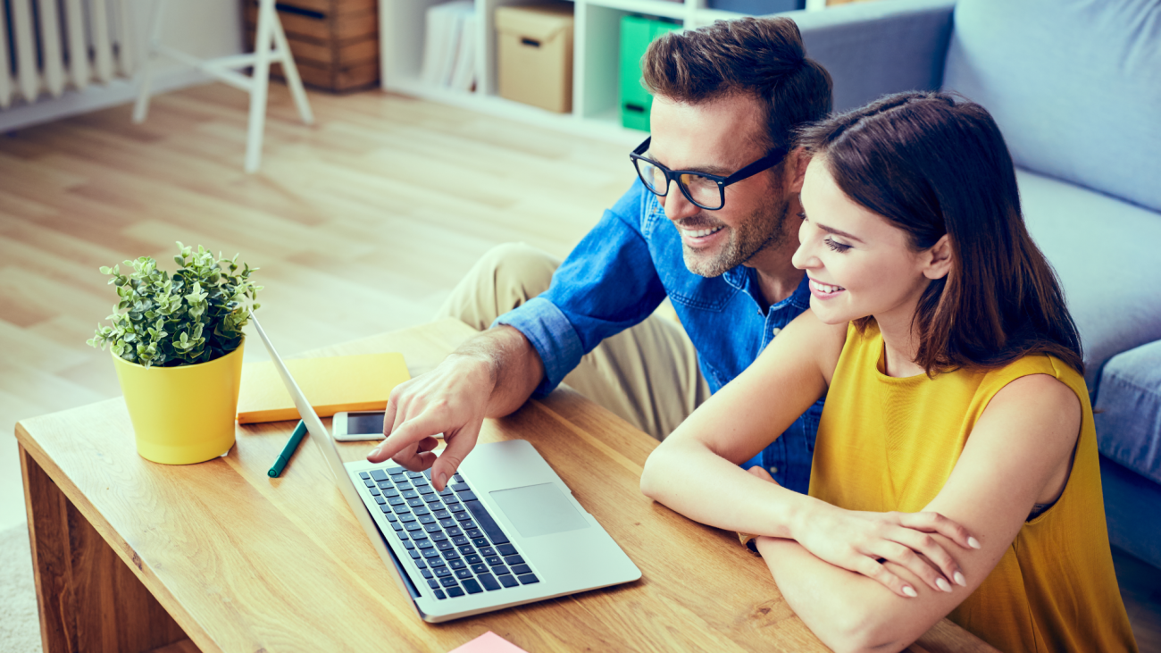 a happy-couple spends time together at home in front of a laptop computer screen, which sits on their coffee table