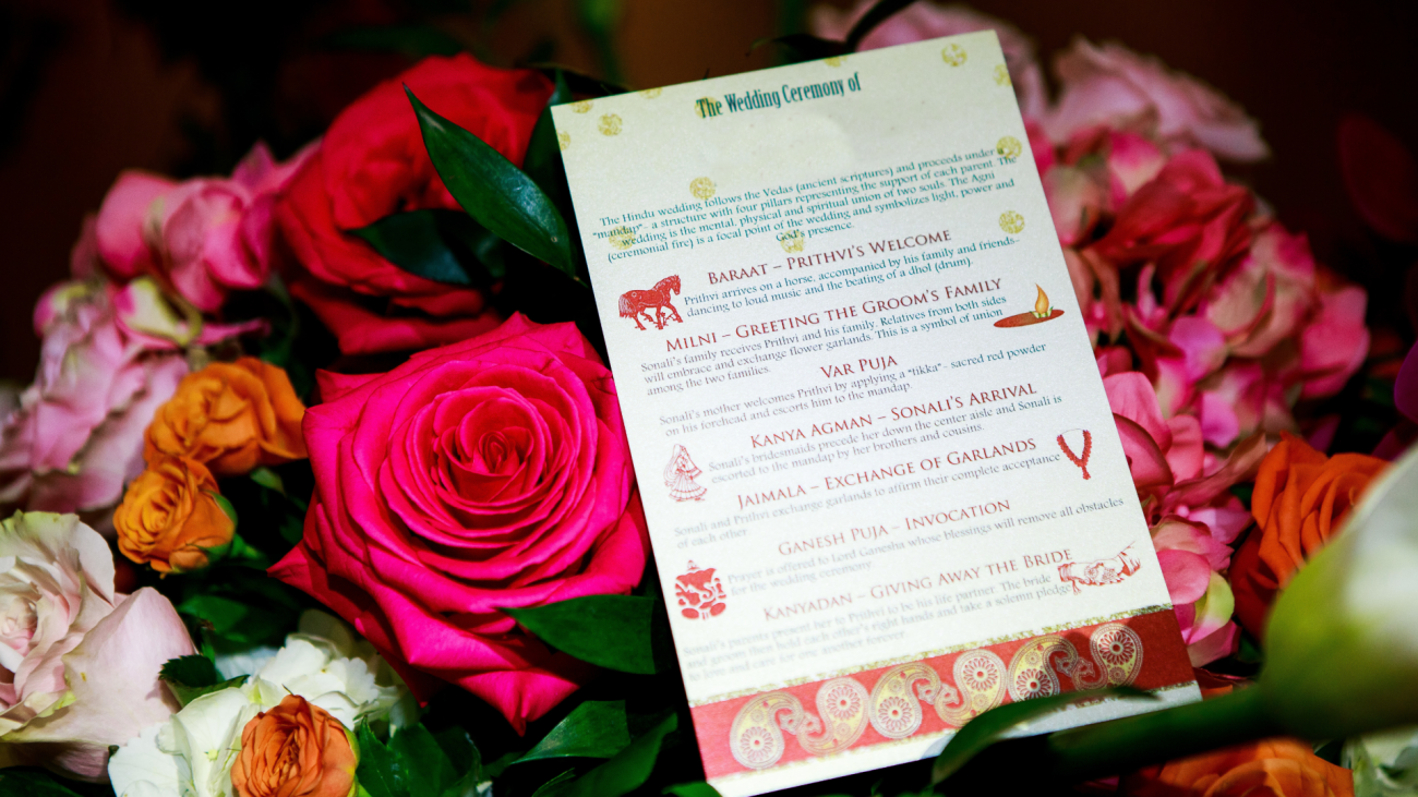 an Indian wedding program card with flowers in the background