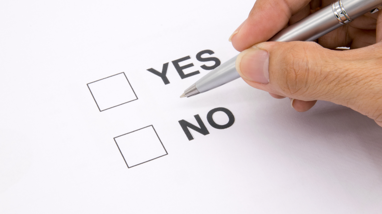 a man’s hand holds a pen over a document and tries to decide between selecting “yes” or “no”