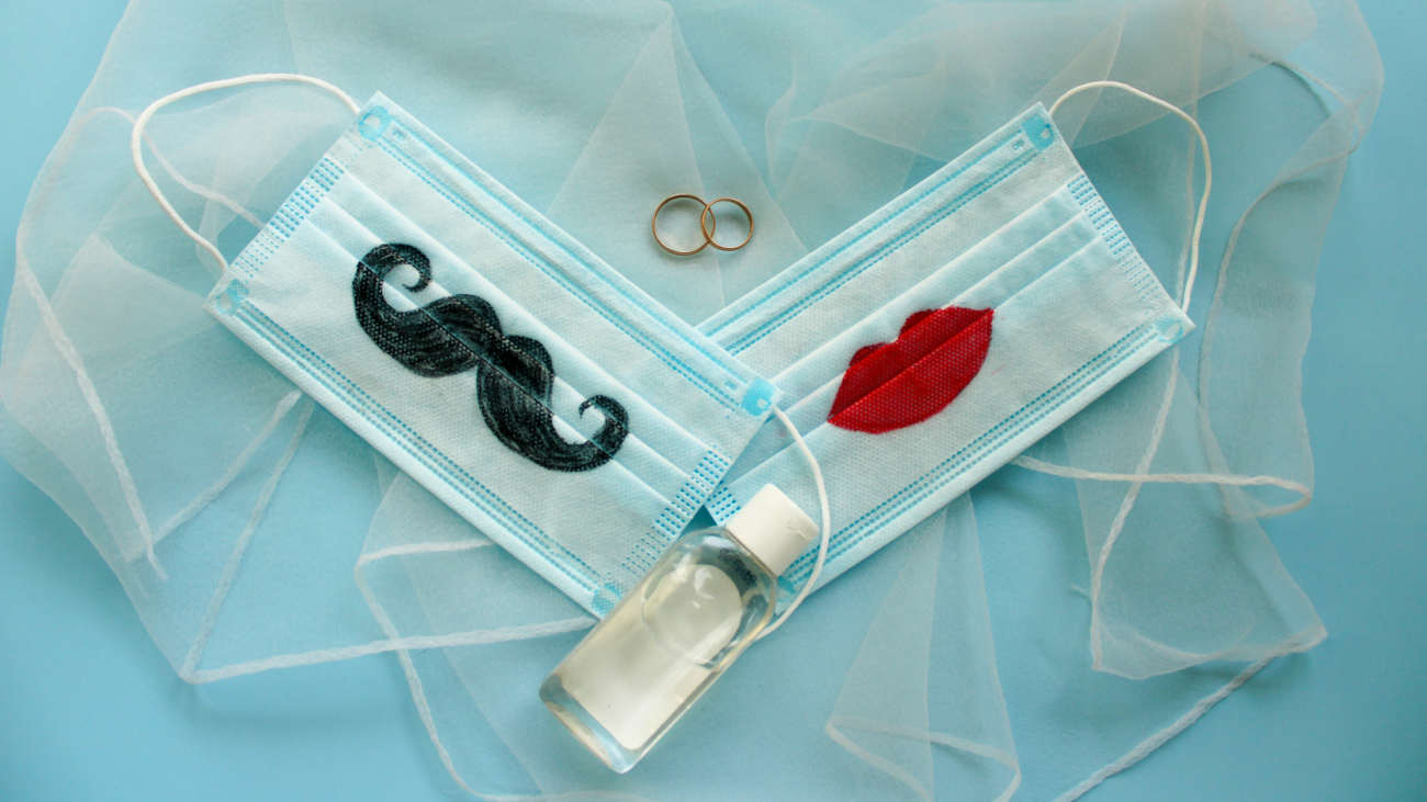two medical face masks painted with a mustache and lips sit next to wedding rings and hand sanitizer