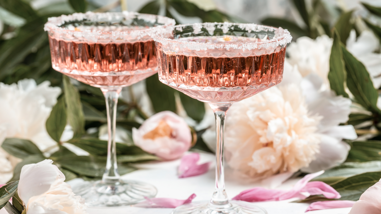 two glasses of rosé wine sit next to peony flowers over a white background