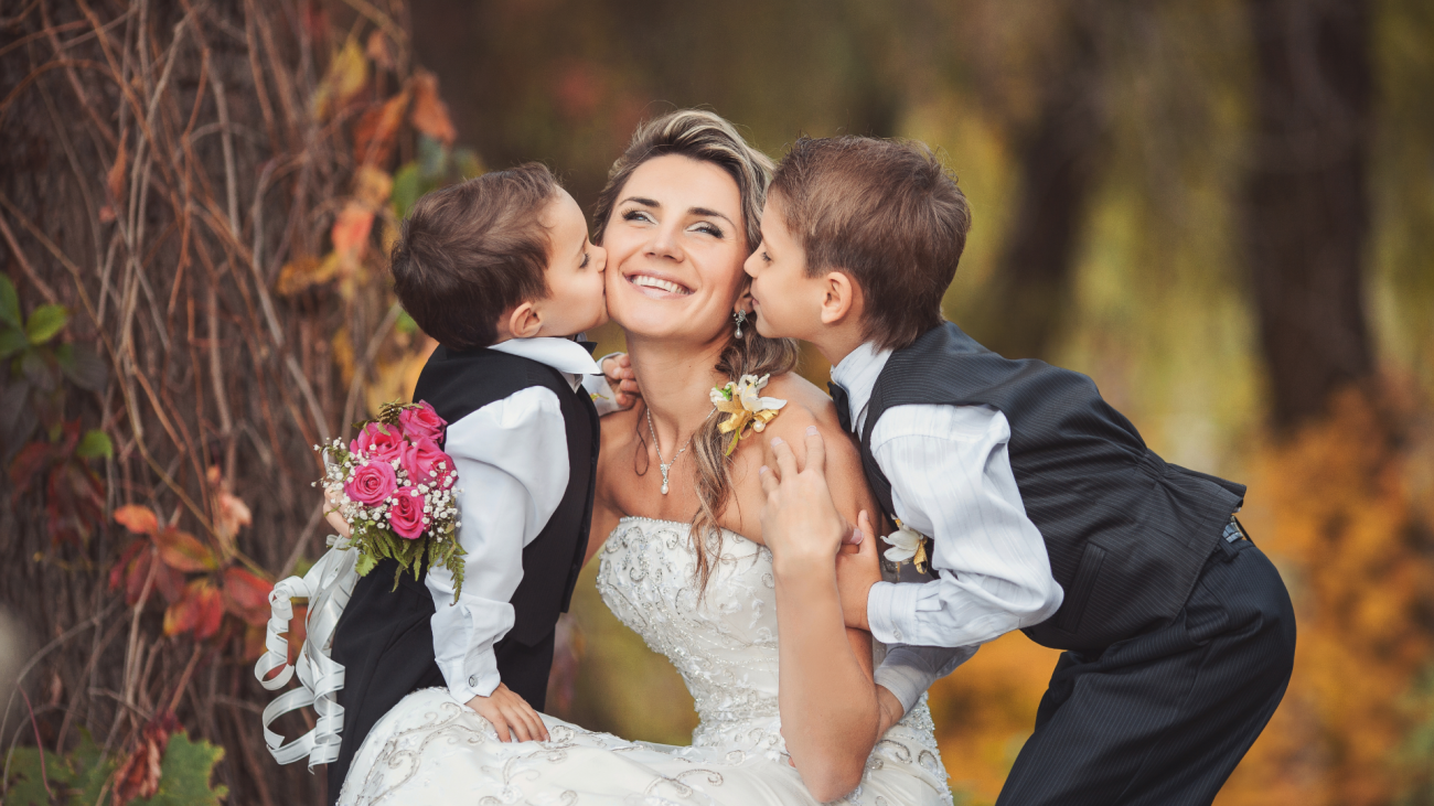 a bride wearing a wedding dress as she kneels on her knees as two children kiss both sides of her cheek