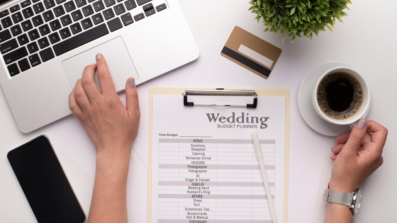 a wedding budget planner checklist sits on a table next to a laptop, a cup of coffee and a smartphone