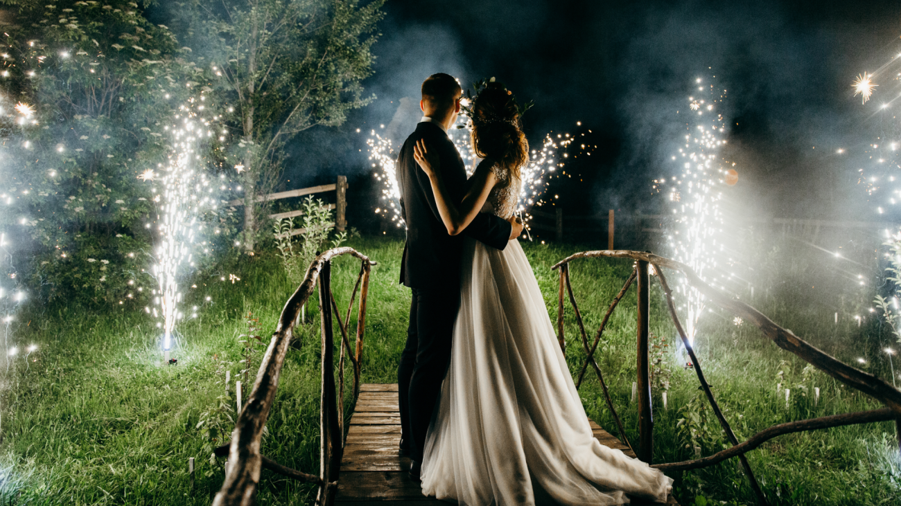 a bride and groom look at fireworks outside on a small bridge