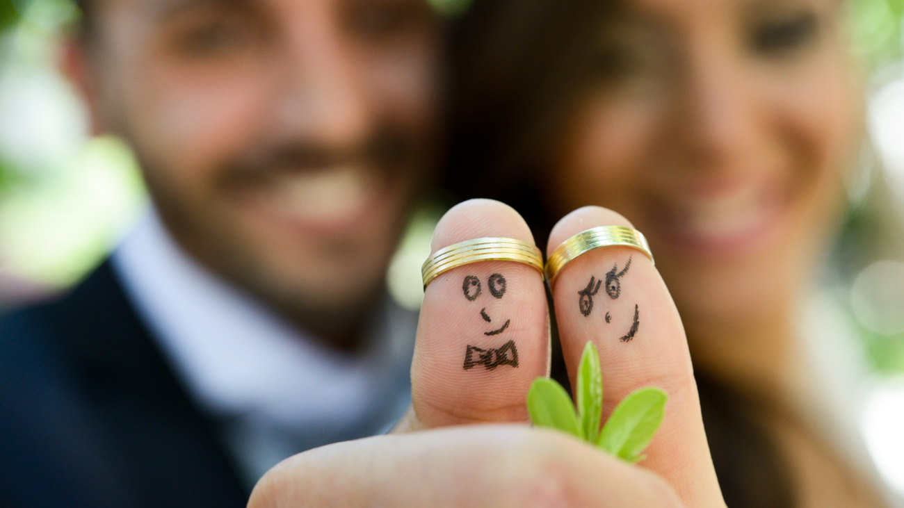 a bride and groom show their thumbs, which sport gold wedding rings and faces drawn on with marker