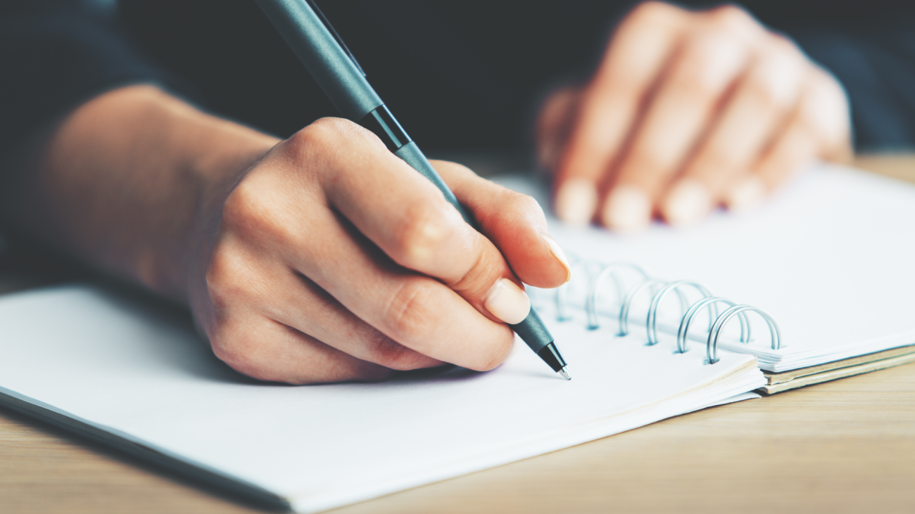 a close-up of a woman's hands writing in a spiral notepad placed on a wooden desktop