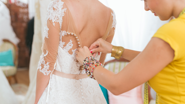 a friend helps the bride fit into her wedding dress by helping to zip up the back