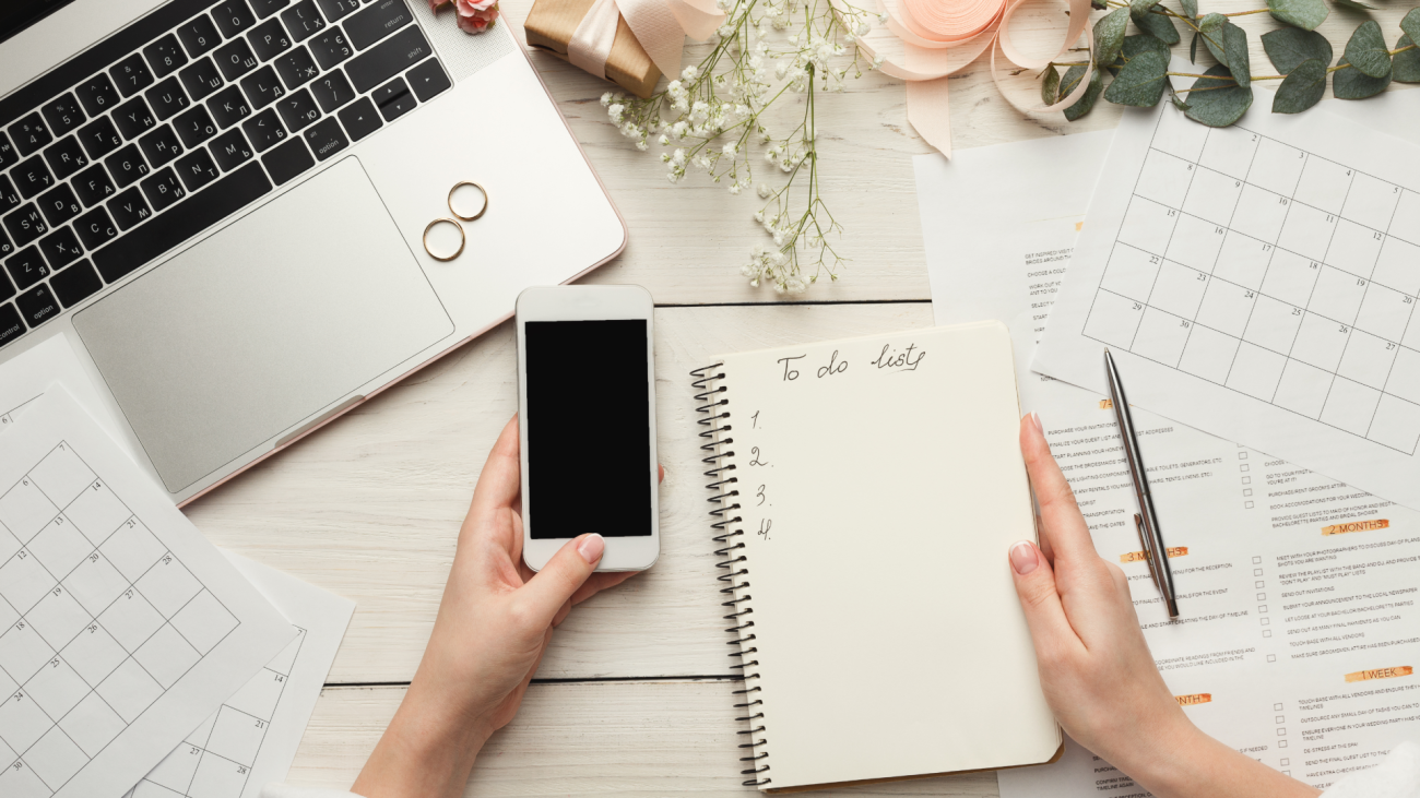 a woman a writes to-do list for her wedding next to laptop computer, smartphone, wedding rings, and flowers