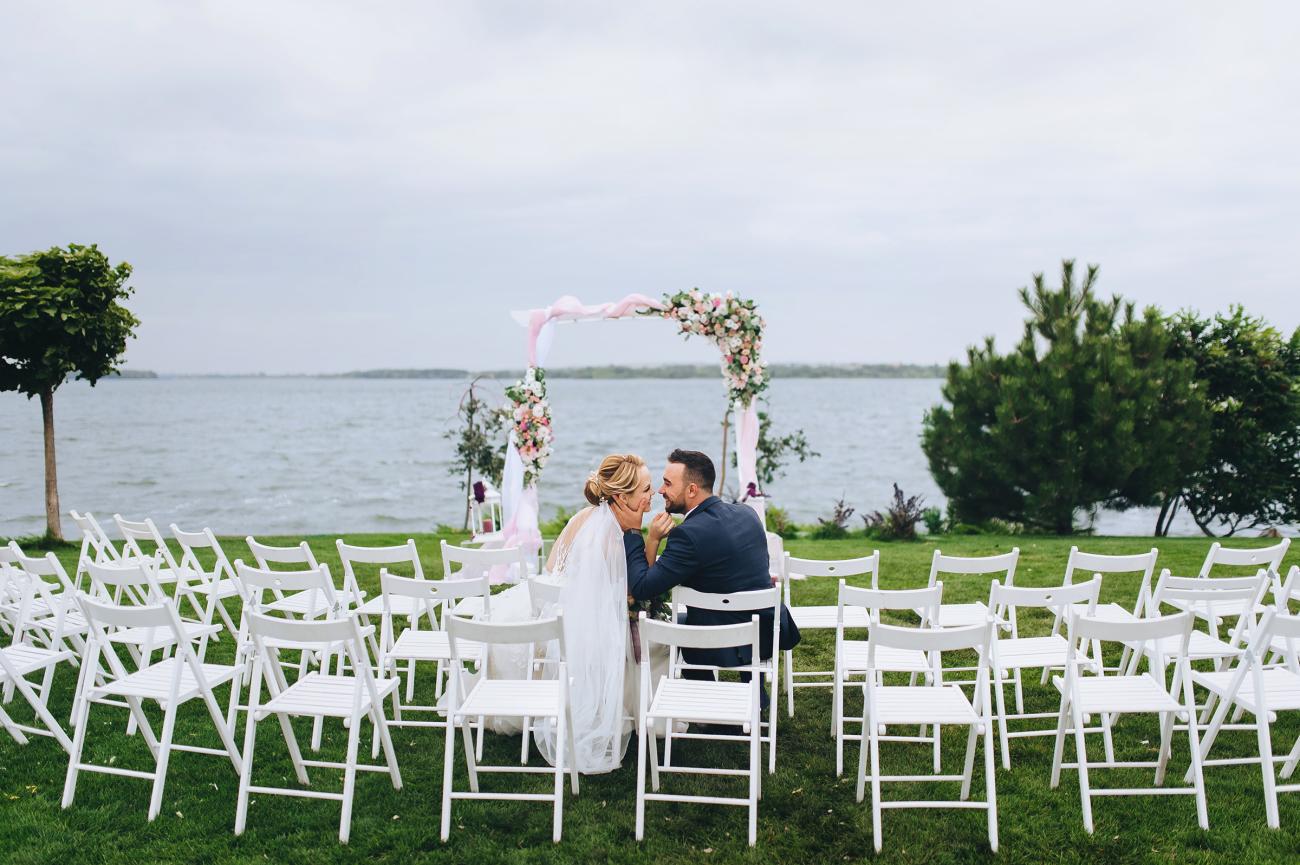 Bride and groom sit on wedding ceremony chairs against the backdrop of an arch and the sea