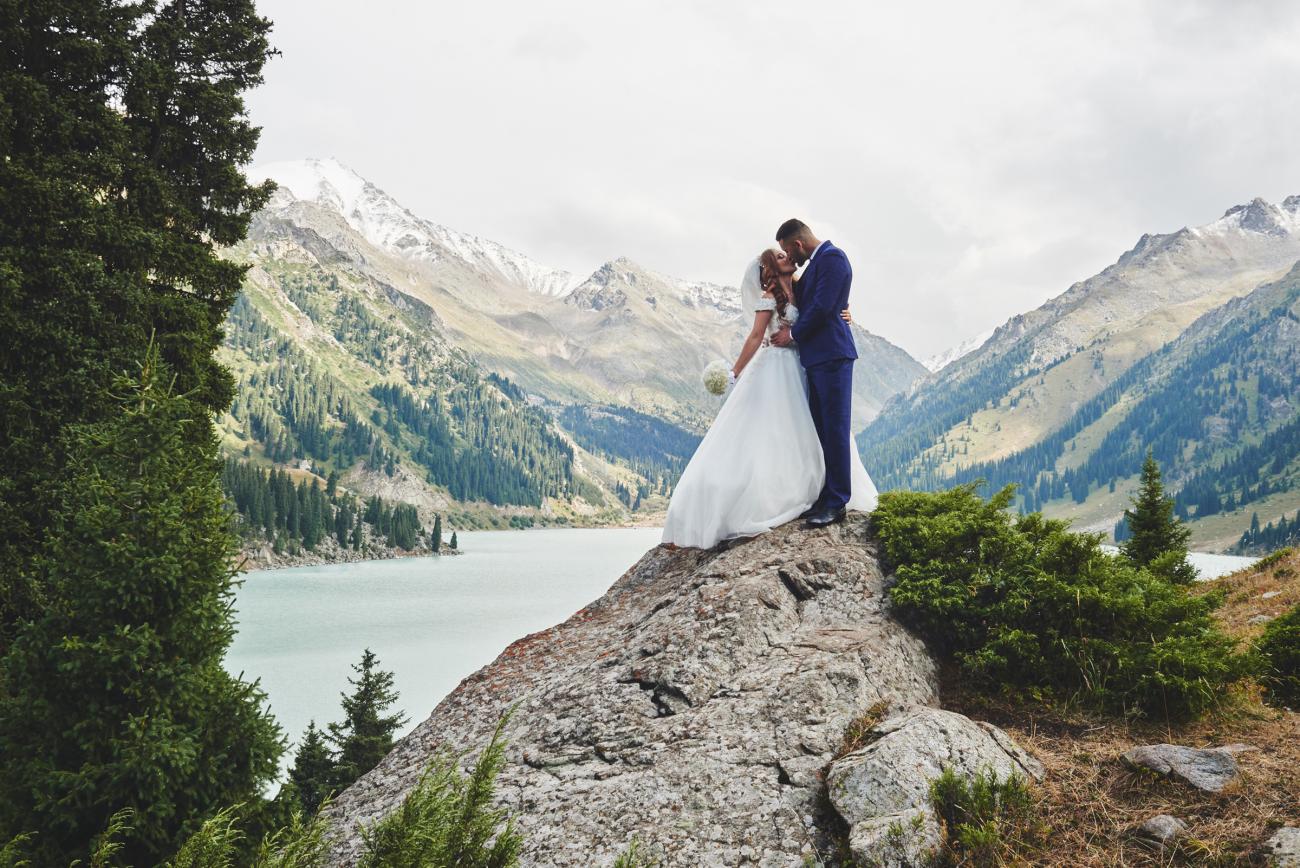 Bride and groom kissing on top of a rock with lake and mountains in the background