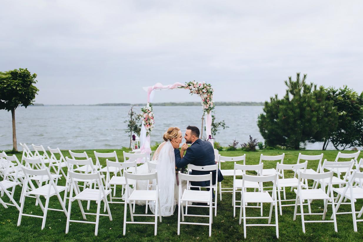 Bride and groom sit on wedding ceremony chairs against the backdrop of an arch and the sea